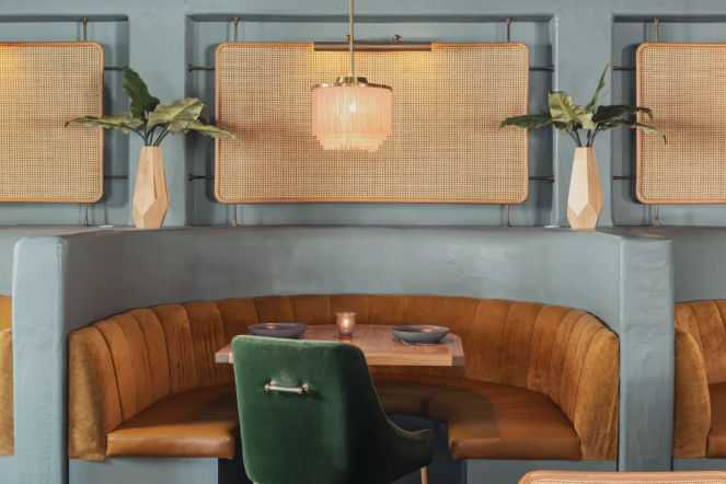 Cozy modern bistro booth with curved caramel banquette, green velvet chair, wooden table for two, rattan wall panels, pendant light, and geometric vases with plants.