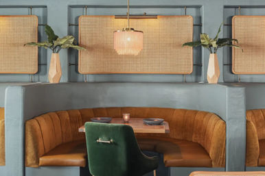 Cozy modern bistro booth with curved caramel banquette, green velvet chair, wooden table for two, rattan wall panels, pendant light, and geometric vases with plants.