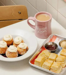White cafe table with a pink ceramic mug of coffee, plate of sugar-coated cream-topped mini donut holes, and a metal tray of flaky buttery biscuit squares with dipping sauces