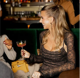 Smiling woman in a lace dress and high ponytail holding a pink cocktail in a coupe glass while chatting with a friend on green velvet seating in a lively cocktail lounge