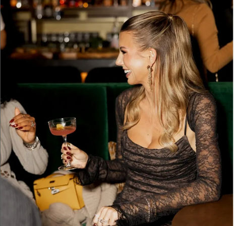 Smiling woman in a lace dress and high ponytail holding a pink cocktail in a coupe glass while chatting with a friend on green velvet seating in a lively cocktail lounge