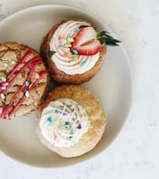 Three decorated donuts on a white plate on a marble countertop — a cinnamon-sugar donut topped with white frosting and rainbow sprinkles, a pink-swirl frosted donut with a halved strawberry, and a cracked cookie-style donut drizzled with pink glaze and sprinkles.