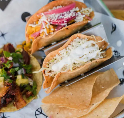 Close-up of a plate of Mexican tacos on a metal rack: two crispy corn tacos with shredded cabbage, crema and cotija cheese, plus an al pastor-style taco topped with pineapple, cilantro and onion, served with tortilla chips.