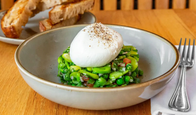 Poached egg crowning a vibrant pea and broad-bean salad with herbs, served in a ceramic bowl with toasted sourdough and a fork on a wooden table.
