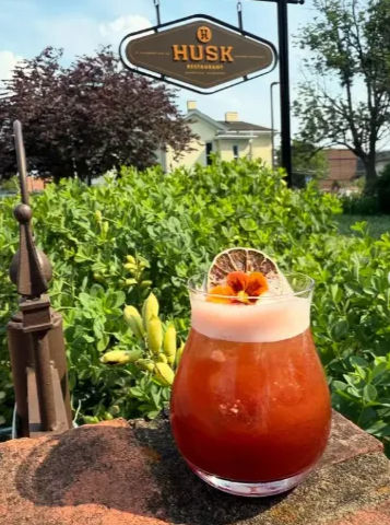 Sunlit red craft cocktail with frothy top, dried citrus wheel and edible flower garnish on a stone ledge, outdoor restaurant patio scene with green shrubs and a hanging sign in the background.