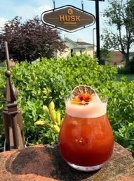 Sunlit red craft cocktail with frothy top, dried citrus wheel and edible flower garnish on a stone ledge, outdoor restaurant patio scene with green shrubs and a hanging sign in the background.
