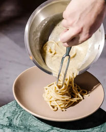 Close-up of a hand plating creamy, pepper-speckled spaghetti into a beige bowl, pouring sauce from a stainless pot with tongs — homemade Italian-style pasta being plated.