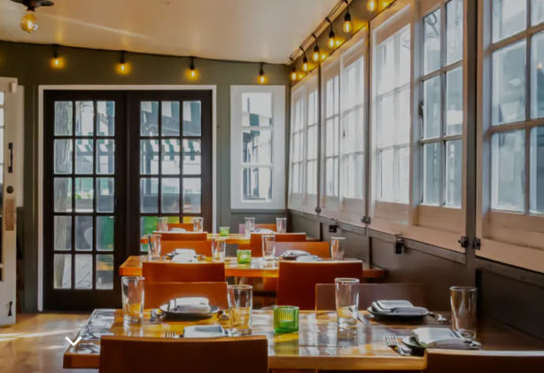 Sunlit restaurant interior with wooden dining tables set with glassware, large paneled windows letting in natural light, warm modern bistro seating and hanging string lights.
