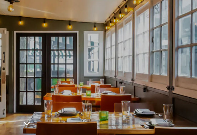 Sunlit restaurant interior with wooden dining tables set with glassware, large paneled windows letting in natural light, warm modern bistro seating and hanging string lights.