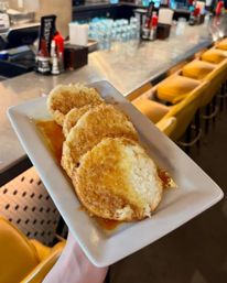 Warm flaky honey-butter biscuits drizzled with syrup on a white rectangular plate held above a diner-style bar lined with yellow barstools