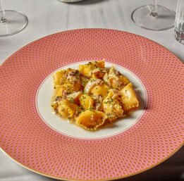 Buttery ravioli in a creamy mushroom sauce topped with chives, plated on a pink patterned rim plate on a white tablecloth with wine glasses — elegant restaurant pasta.