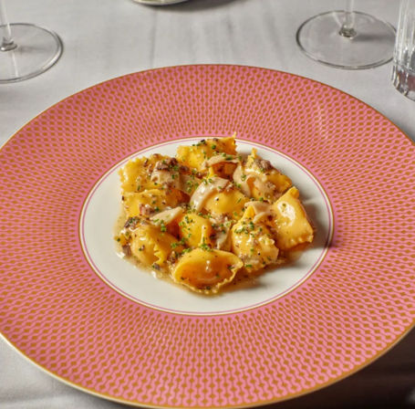 Buttery ravioli in a creamy mushroom sauce topped with chives, plated on a pink patterned rim plate on a white tablecloth with wine glasses — elegant restaurant pasta.