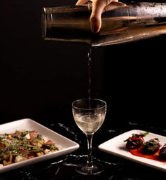 Hand pouring a clear cocktail from a metal shaker into a coupe glass on a black marble bar, flanked by two small appetizer plates for a chic cocktail bar presentation.