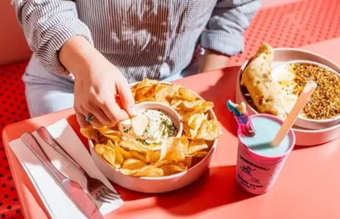 A hand dipping a chip into a herby cream dip surrounded by crispy kettle chips, next to a savory pastry with grains and a pastel blue milkshake on a bright pink cafe table.