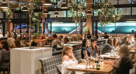 Busy modern urban restaurant interior at night with diners at wooden communal tables and cozy booths, hanging pendant lights, indoor trees, and large glass windows overlooking city buildings.