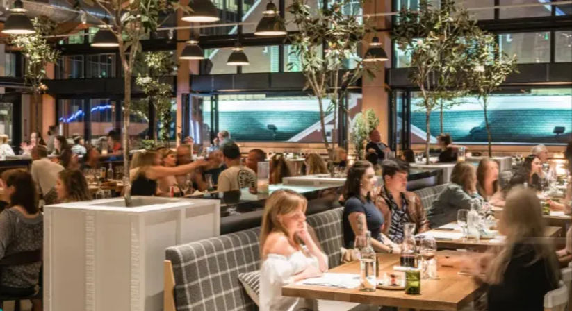 Busy modern urban restaurant interior at night with diners at wooden communal tables and cozy booths, hanging pendant lights, indoor trees, and large glass windows overlooking city buildings.