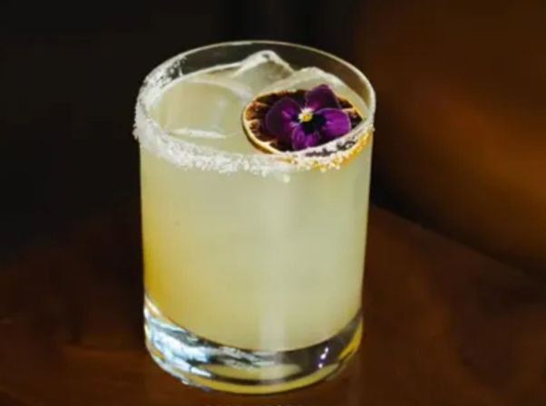 Salt-rimmed margarita-style cocktail in a short rocks glass with ice, pale citrus liquid, dried citrus wheel and purple edible flower garnish on a wooden bar