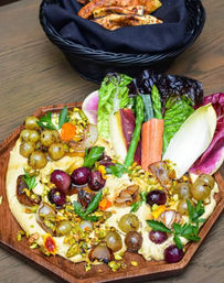 Mediterranean-style hummus platter on a wooden board topped with roasted green and red grapes, caramelized onions, chopped pistachios and parsley, served with colorful romaine, radicchio, carrot sticks and asparagus and a basket of pita