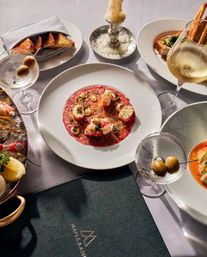 Candlelit fine-dining spread: vibrant red carpaccio topped with shrimp and roe on a white plate, martini with olives, glass of white wine, toasted bread, seafood platter and bowl of soup.