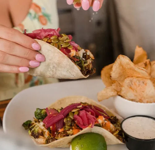 Hand with pink nails squeezing lime over roasted vegetable tacos topped with pickled red onions and pumpkin seeds, served with kettle chips and creamy dip