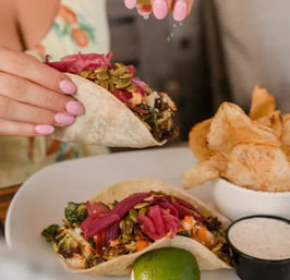 Hand with pink nails squeezing lime over roasted vegetable tacos topped with pickled red onions and pumpkin seeds, served with kettle chips and creamy dip