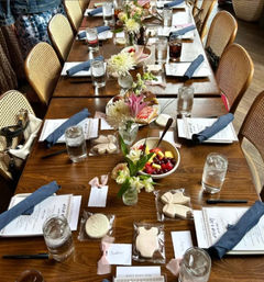 Long wooden table in a cozy indoor restaurant set for a baby shower brunch with rattan chairs, blue cloth napkins, glass water tumblers, small floral centerpieces, bowls of fresh fruit, printed menus, name cards and individually wrapped baby-themed cookies.