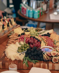 Artisan charcuterie grazing board on a wooden serving board with assorted cheeses, cured meats, red and green grapes, blackberries, fresh figs, green olives, crackers and a small dip, styled on a buffet table at an indoor event.