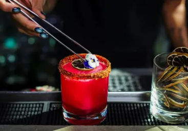 Bright crimson craft cocktail in a short glass with a chili-sugar rim being garnished with an edible pansy using metal tweezers, a glass of dried citrus slices sits beside it on a dimly lit bar.