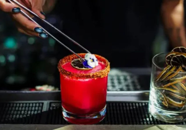 Bright crimson craft cocktail in a short glass with a chili-sugar rim being garnished with an edible pansy using metal tweezers, a glass of dried citrus slices sits beside it on a dimly lit bar.