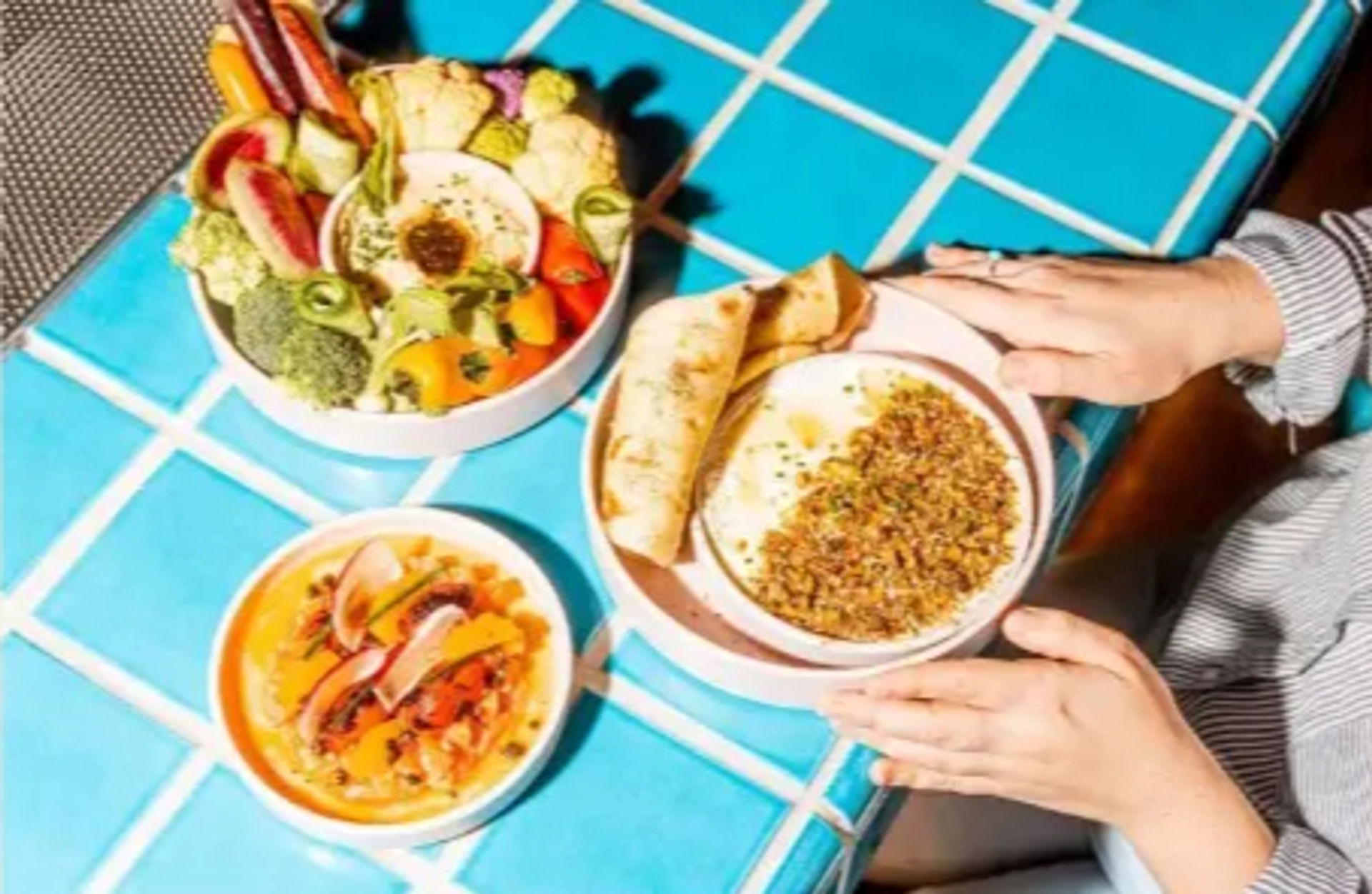 Mediterranean-style hummus bowl topped with grains and pita, colorful vegetable and salad bowls on a bright blue tiled cafe table with hands reaching in