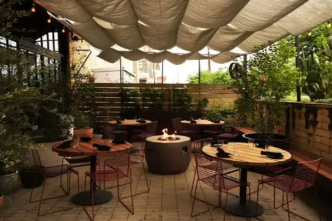 Cozy urban outdoor dining patio with round wooden tables, red wire chairs, a central fire pit, fabric canopy overhead and lush potted greenery