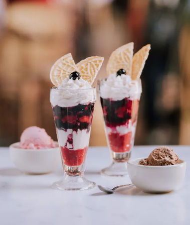 Two layered berry parfaits in tall glasses with whipped cream, wafer cookies and cherries, flanked by bowls of strawberry and chocolate ice cream on a white table — café-style dessert