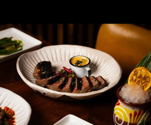 Sliced medium-rare steak fanned in a white textured bowl with charred crust, paired with a small cup of creamy yellow dipping sauce and pickled garnish on a wooden table, with a tiki-style cocktail topped with crushed ice and a dried citrus wheel nearby.