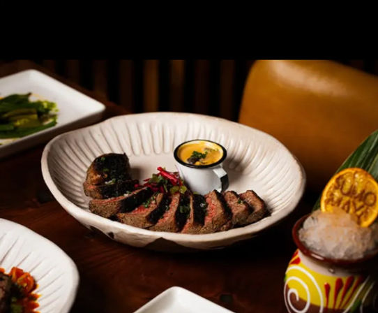 Sliced medium-rare steak fanned in a white textured bowl with charred crust, paired with a small cup of creamy yellow dipping sauce and pickled garnish on a wooden table, with a tiki-style cocktail topped with crushed ice and a dried citrus wheel nearby.