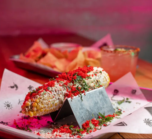 Elote (Mexican street corn) on a metal stand topped with crema, cotija cheese, crushed red chips and chopped cilantro, served with tortilla chips and a margarita in a pink-lit restaurant setting