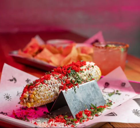 Elote (Mexican street corn) on a metal stand topped with crema, cotija cheese, crushed red chips and chopped cilantro, served with tortilla chips and a margarita in a pink-lit restaurant setting