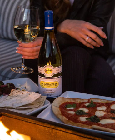 Cozy outdoor patio scene with a bottle of Chardonnay and a glass of white wine held by a person, margherita pizza and flatbreads on plates beside a glowing fire pit