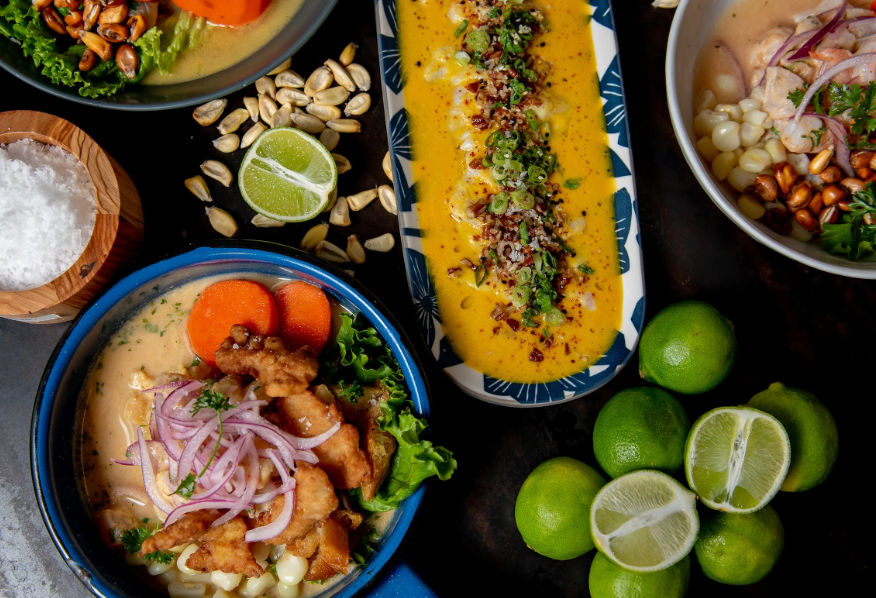 Overhead shot of Peruvian-style seafood bowls and a long dish of bright ají amarillo sauce, garnished with fried fish, pickled red onions, cancha corn, peanuts and halved limes on a dark tabletop.