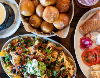 Top-down restaurant brunch spread: loaded nachos with guacamole, sour cream, black beans and pico, basket of salted pretzel rolls, grilled salmon with sliced tomatoes and pickled onions, and a small pot of cheese dip.