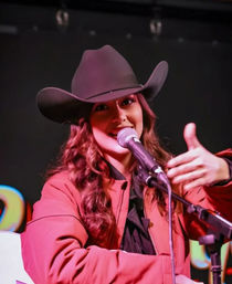 Country singer in a black cowboy hat and red jacket smiling into a microphone on stage, making a thumbs-up gesture during a live country music performance