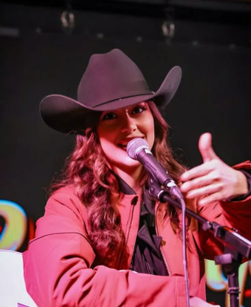 Country singer in a black cowboy hat and red jacket smiling into a microphone on stage, making a thumbs-up gesture during a live country music performance