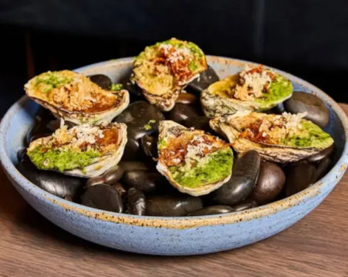 Six oysters on the half shell topped with green herb sauce and crispy breadcrumbs, arranged on black stones in a blue ceramic bowl — seafood appetizer, restaurant-style plating.
