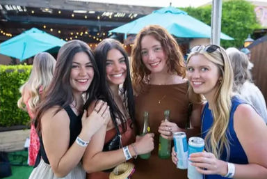 Four smiling young women holding bottles and cans on a sunny outdoor patio with teal umbrellas and string lights — casual summer friends gathering.