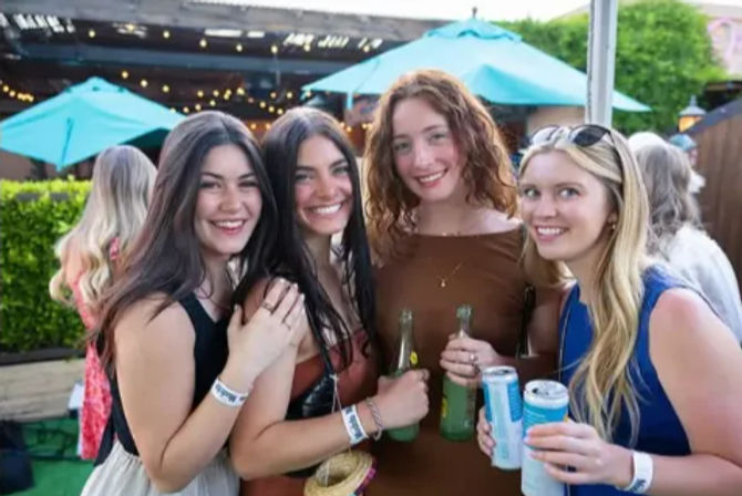 Four smiling young women holding bottles and cans on a sunny outdoor patio with teal umbrellas and string lights — casual summer friends gathering.