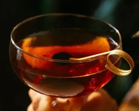 Close-up of an amber cocktail in a coupe glass with a dark cherry and curled citrus peel garnish at a dim cocktail bar.