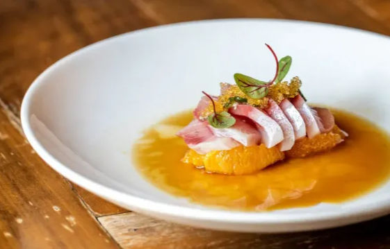 Gourmet seafood appetizer: thinly sliced raw fish on citrus segments with zesty orange sauce, microgreens and fish roe, served on a white plate on a wooden table.
