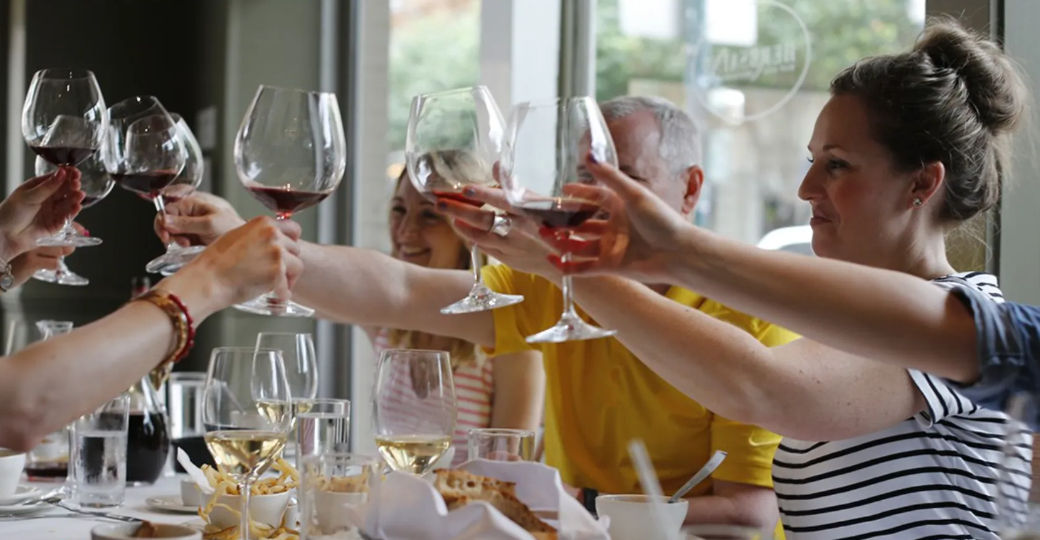 Cheerful group toasting with red wine glasses over a casual restaurant table set with plates, white wine and snacks