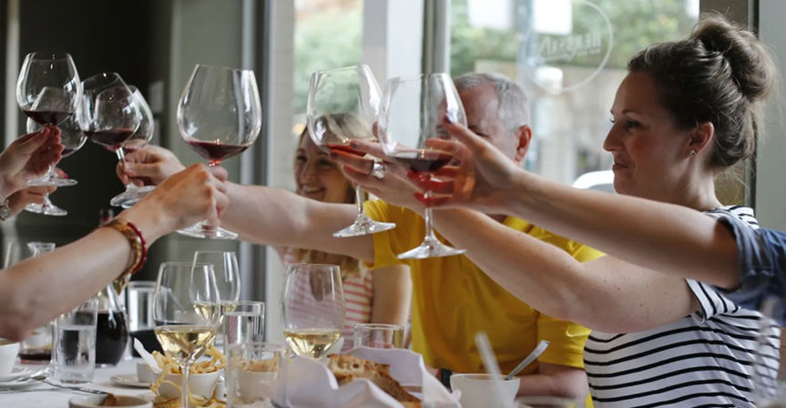Cheerful group toasting with red wine glasses over a casual restaurant table set with plates, white wine and snacks