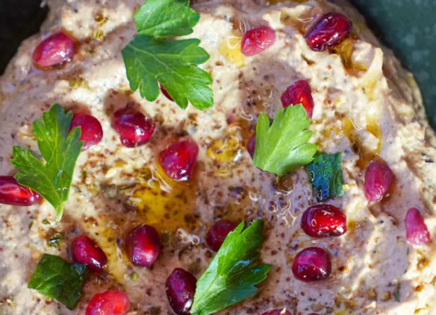 Close-up overhead of creamy hummus in a bowl drizzled with olive oil, sprinkled with za'atar and ruby pomegranate seeds, and garnished with fresh parsley — vibrant Mediterranean dip.