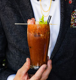 Tall Bloody Mary-style spicy tomato cocktail with chili-salt rim, salami-and-cheese skewer, celery stalk and black straw, held in hand against a dark jacket background.